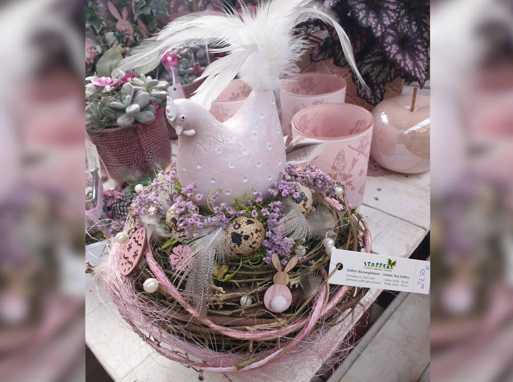 Rosa Tischdekoration. In einem Nest sitzt eine Hühnerfigur, umgeben von lila Blüten und Wachteleiern.