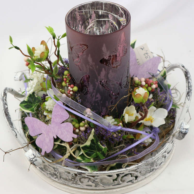  Ein weißes Dekotablett mit einem lila Kerzenglas; dekoriert mit zahlreichen Kunstblüten und -beeren. 