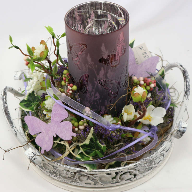 Ein weißes Dekotablett mit einem lila Kerzenglas; dekoriert mit zahlreichen Kunstblüten und -beeren.