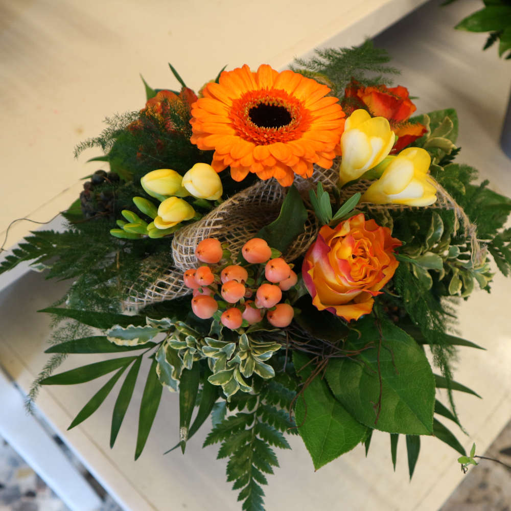 Ein kleiner, kompakt gebundener Blumenstrauß, bestehend aus orangen Gerbera und orange-gelben Rosenblüten; ergänzt um gelbe Blüten.